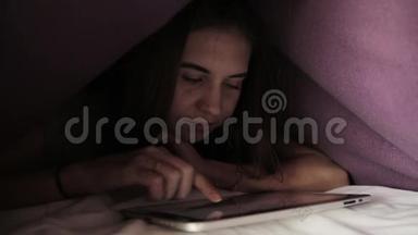 年轻女孩躲在羽绒被下，在黑暗中使用PC平板设备。 青少年晚上在数字平板电脑上玩。